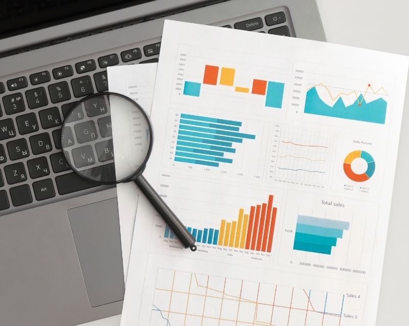 Financial charts and analytical reports reviewed with a magnifying glass, reflecting forensic analysis and business intelligence assessment.